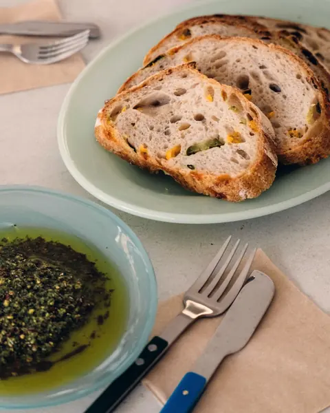 Scheiben von Sauerteigbrot auf einem hellgrünen Teller, davor eine Schale Olivenöl mit Kräutern