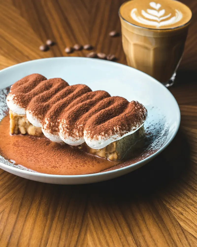 Tiramisù mit Kakao bestäubt auf einem hellen Teller, dahinter ein Glas Milchkaffee